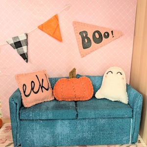 Könnte beinhalten: Ein Miniatur-Sofa in Türkis mit Kissen in Kürbis- und Geisterform und einem Banner mit der Aufschrift "BOO!". Die Szene ist mit einem kissenförmigen Kürbis, einem kissenförmigen Geist und einem Banner mit dem Wort "BOO!" dekoriert.