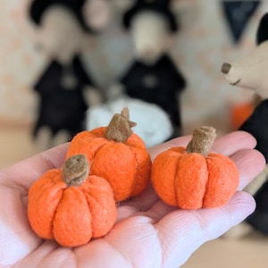 May include: Three small, orange felt pumpkins with brown stems are held in a hand. The pumpkins are in varying sizes and have a ribbed texture. The background includes blurred Halloween decorations, suggesting a seasonal theme.