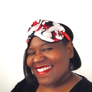 May include: A woman wearing a black sleep mask with a red and white crane pattern. The mask has an elastic band.