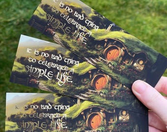 May include: Three bookmarks with a green, mossy background featuring a hobbit hole with a round wooden door. The bookmarks have gold lettering that reads "It is no bad thing to celebrate a simple life."