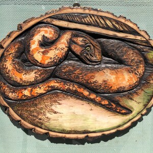May include: A wood carving of a snake coiled on a tree trunk slice. The snake is carved in detail and painted with orange and brown tones.