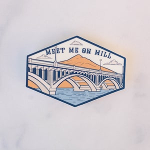 May include: A sticker with a blue and white illustration of a bridge over a body of water. The text "MEET ME ON MILL" is written in a white font on a blue background.