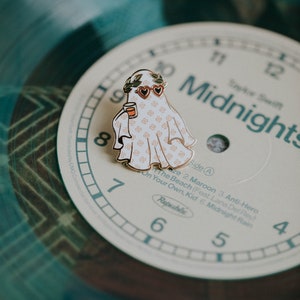 May include: A gold-colored enamel pin featuring a ghost wearing heart-shaped sunglasses and a wreath of leaves. The ghost is holding a small cup and is patterned with a white and pink design. The pin is on top of a vinyl record with the text "Midnights" printed on it.
