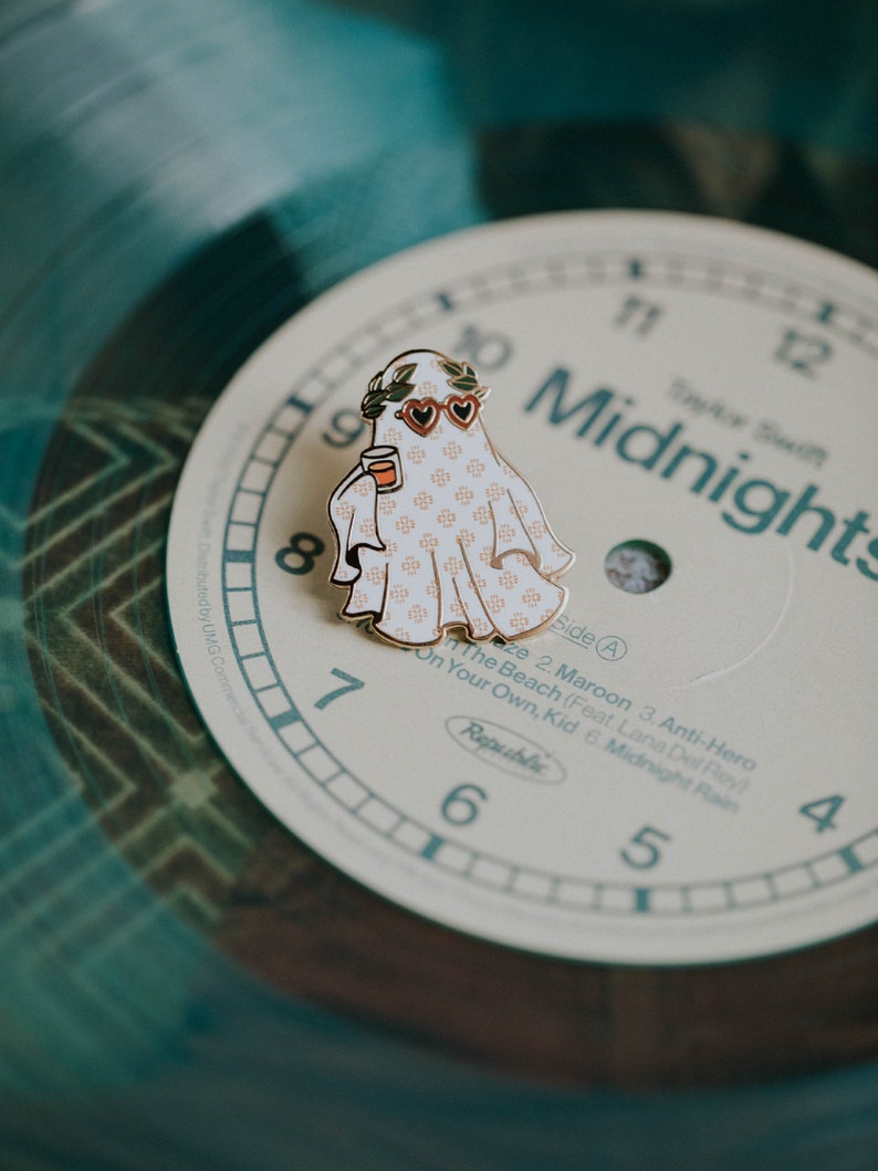 May include: A gold-colored enamel pin featuring a ghost wearing heart-shaped sunglasses and a wreath of leaves. The ghost is holding a small cup and is patterned with a white and pink design. The pin is on top of a vinyl record with the text "Midnights" printed on it.