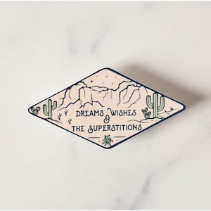 May include: A diamond-shaped sticker featuring a desert landscape with cacti, mountains, and stars. The sticker has a cream background with dark blue and green accents. The text "DREAMS WISHES & THE SUPERSTITIONS" is displayed in a dark blue font.