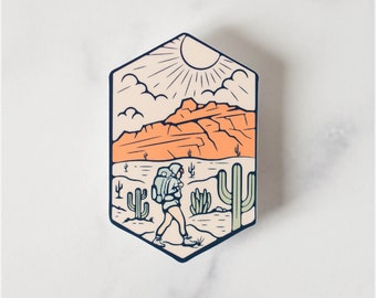 May include: A hexagonal sticker featuring a desert landscape with a hiker. The design includes a sun, clouds, mountains, and cacti in shades of orange, beige, and green. The hiker is wearing a backpack and walking through the desert.