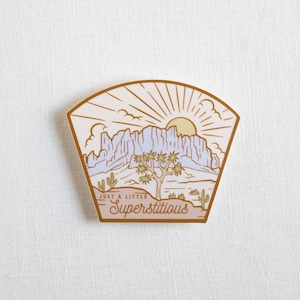 May include: A beige and brown sticker with a desert landscape illustration. The sticker features a mountain range, a Joshua tree, and a sun with rays. The text "Just a little Superstitious" is written in a brown script font.