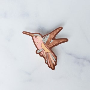 May include: A sticker of a hummingbird with brown, pink, and blue wings in flight. The hummingbird is facing to the right.