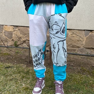 Op de afbeelding: Blauwe, witte en lichtblauwe sweatpants met een cartoonfiguur bedrukt op het rechterbeen. Het figuur is een blauwe alien met grote ogen en een brede glimlach. Het linkerbeen heeft de woorden "Ohana" meerdere keren in het zwart herhaald. De sweatpants worden gedragen door iemand die paarse high-top sneakers draagt.