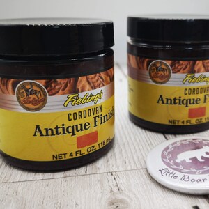 May include: Two 4-ounce bottles of Fiebing's Cordovan Antique Finish leather dye. The bottles are yellow with brown text and a brown leather design. The bottles are sitting on a white wooden surface.