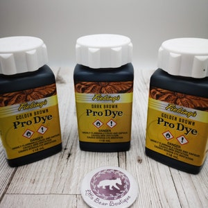 May include: Three bottles of Fiebing's Pro Dye leather dye. The bottles are labeled with the colors Golden Brown and Dark Brown. Each bottle contains 118 ml of dye. The bottles are sitting on a wooden surface.