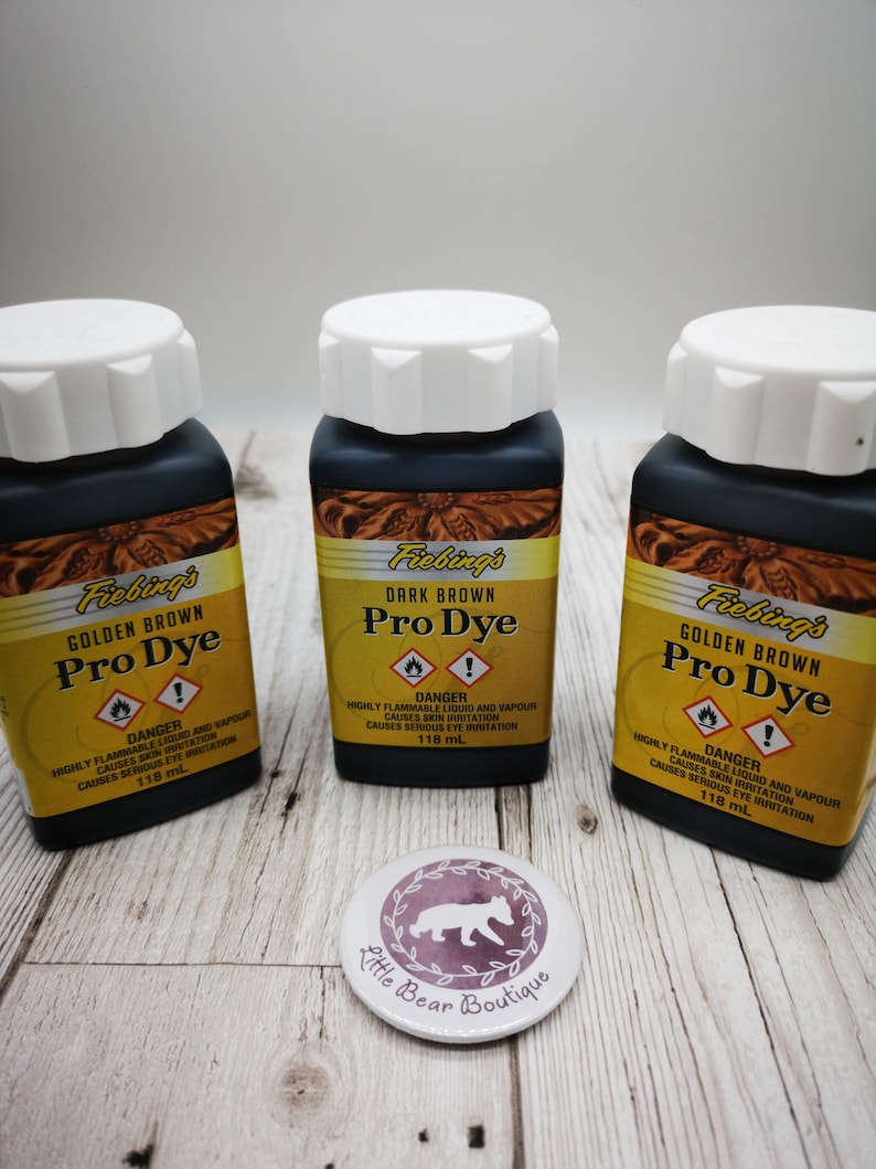 May include: Three bottles of Fiebing's Pro Dye leather dye. The bottles are labeled with the colors Golden Brown and Dark Brown. Each bottle contains 118 ml of dye. The bottles are sitting on a wooden surface.