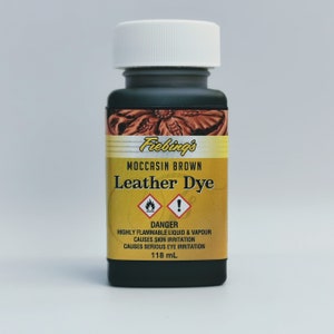 May include: A small bottle of Fiebing's Leather Dye in Moccasin Brown. The bottle is black with a yellow label and a white cap. The label has a warning that the liquid is highly flammable and causes skin and eye irritation. The bottle contains 118 ml of dye.