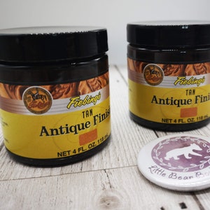May include: Two brown glass jars with black lids containing Fiebings Tan Antique Finish leather dye. Each jar has a yellow label with the product name and size: 4 fl. oz. (118 ml).
