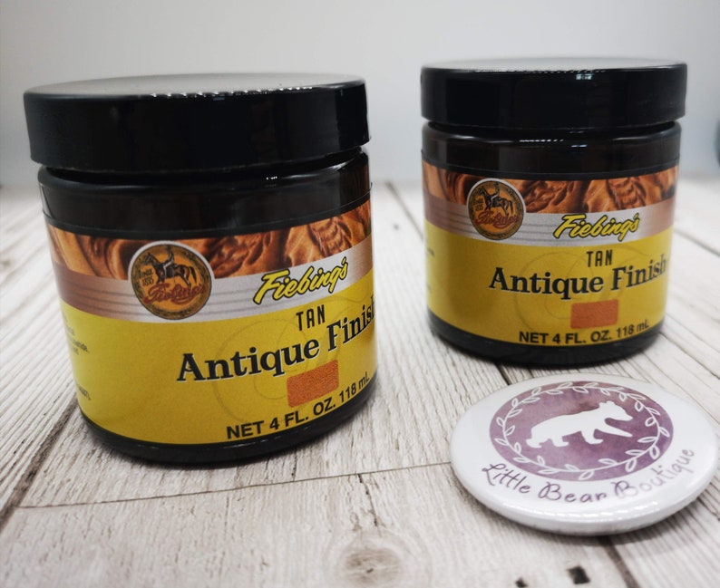 May include: Two brown glass jars with black lids containing Fiebings Tan Antique Finish leather dye. Each jar has a yellow label with the product name and size: 4 fl. oz. (118 ml).