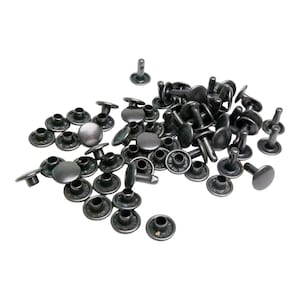 May include: A pile of dark grey metal rivets, each with a flat, round head and a post. The rivets are scattered on a white background, with some standing upright and others lying flat. These fasteners are commonly used in leathercraft and garment construction.