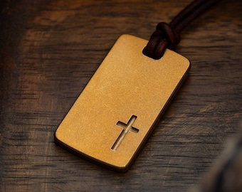 May include: A gold-colored rectangular pendant with a cut-out cross design, suspended from a brown leather cord. The pendant rests on a dark wooden surface, with a rope and a dark wooden tray in the background.
