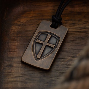 May include: A bronze pendant necklace with a shield design featuring a cross. The pendant is on a wooden surface.