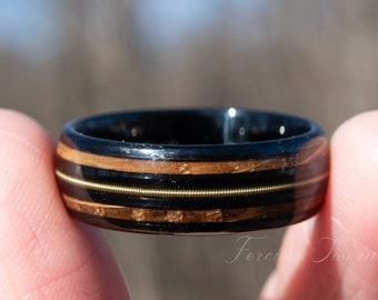 May include: A black ceramic ring with a wood inlay and two thin gold bands. The ring is being held by a finger.