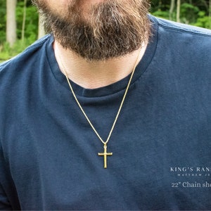 May include: A gold-colored cross pendant on a gold chain. The necklace is worn over a dark blue t-shirt. The text "KING'S RANSOM Matthew 20:28" and "22" Chain shown" is visible in the lower right corner.