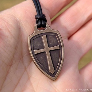 May include: Antiqued bronze shield pendant with a cross design, hanging from a black leather cord.  King's Ransom Matthew 20:28.