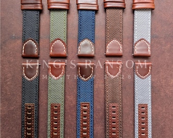 May include: A collection of five watch straps in various colors: black, olive green, navy blue, brown, and gray. Each strap features a brown leather accent and a black metal buckle. The straps are arranged vertically against a brown background.