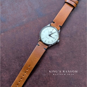 Pode incluir: Um relógio de pulso prateado com mostrador branco e ponteiros pretos, com uma bracelete de couro castanho. O relógio tem um design clássico. O texto "KING'S RANSOM" e "MATTHEW 20:28" estão impressos abaixo.