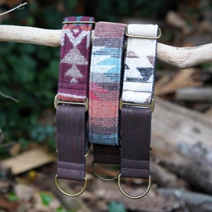 Op de afbeelding: Drie hondenhalsbanden met verschillende geometrische patronen in tinten bruin, rood, wit en blauw. De halsbanden zijn gemaakt van stof en hebben bruine leren banden met messing gespen.