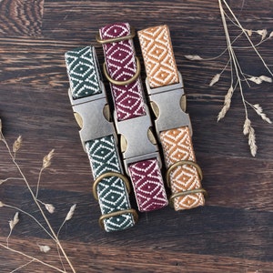 Puede incluir: Tres collares para perros con patrones geométricos en verde, burdeos y beige, cada uno con hebilla y anilla metálica. Los collares están dispuestos sobre una superficie de madera oscura, con acentos de hierba seca.