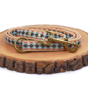 Puede incluir: Una correa para perros con un patrón tejido en beige, naranja, gris y azul claro. La correa tiene un cierre de color latón y un anillo a juego. La correa está sobre una rodaja de madera.