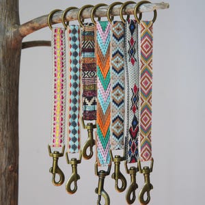 May include: Seven patterned woven straps with brass-toned metal clasps and rings. Each strap displays a different geometric design in colours such as pink, blue, orange, and brown. The straps are suspended from a wooden branch.