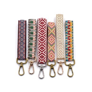 Puede incluir: Conjunto de seis llaveros coloridos con patrones geométricos y clips giratorios dorados. Los llaveros están hechos de tejido tejido y presentan una variedad de colores, incluyendo rojo, azul, verde, amarillo y naranja.