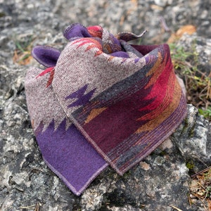 May include: A folded, patterned bandana in shades of purple, red, brown, and gray, resting on a gray rock. The bandana features a geometric design with a purple border and a textured weave.