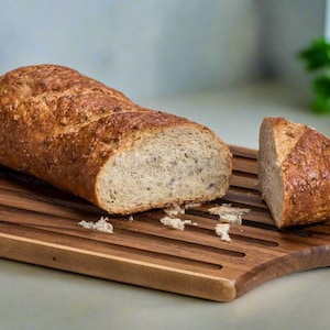 May include: A loaf of freshly baked bread, partially sliced, rests on a wooden cutting board. The bread has a golden-brown crust and a light interior. A slice of bread is cut and placed next to the loaf. An olive oil bottle is in the background.