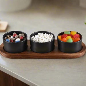 May include: Three black ceramic bowls filled with colorful candies, white candy, and chocolate candies, sitting on a wooden tray.