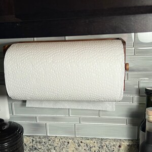 May include: A wooden paper towel holder mounted on a wall with a roll of white paper towels. The holder is made of dark wood and has a simple design. The paper towels are hanging from the holder and are ready to be used.