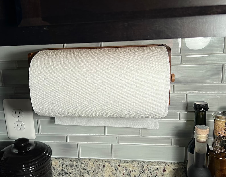 May include: A wooden paper towel holder mounted on a wall with a roll of white paper towels. The holder is made of dark wood and has a simple design. The paper towels are hanging from the holder and are ready to be used.