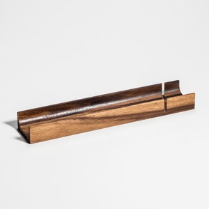 May include: A rectangular wooden desk organizer with a curved interior and a slot. The organizer is made of dark brown and light brown wood, with a smooth finish. The design is minimalist and modern, suitable for office or home use.