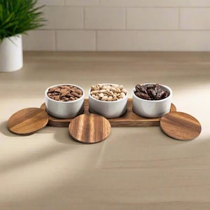 May include: Three white ceramic bowls filled with almonds, pistachios, and dates, sitting on a wooden serving tray with three matching wooden lids.