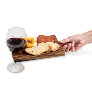 May include: A wooden serving board with a handle, featuring compartments for cheese, crackers, and salami. A glass of red wine is next to the board.