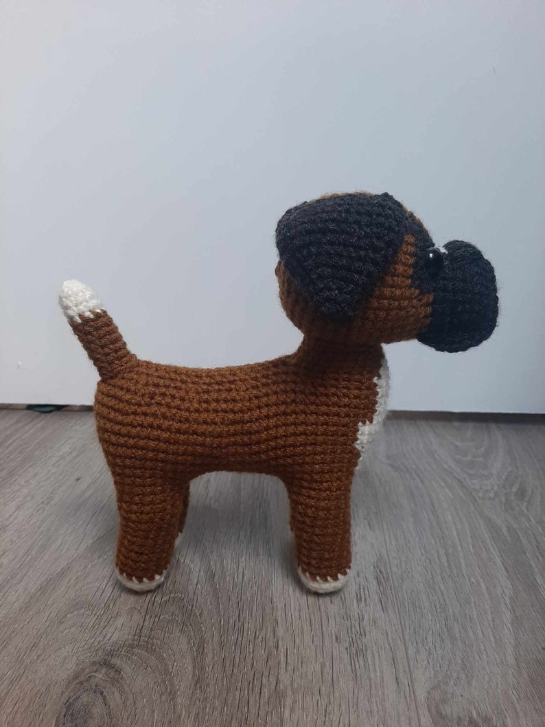 Op de afbeelding: Een gehaakt boxerhondje speelgoed in bruin en wit. De hond staat op een houten vloer.
