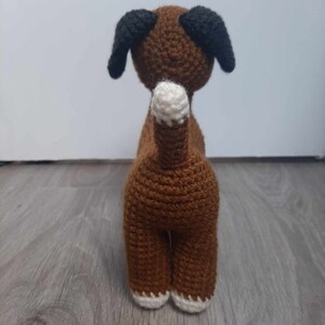Op de afbeelding: Bruin gehaakt hondenspeelgoed met zwarte oren en witte voeten. De hond staat op een houten vloer.