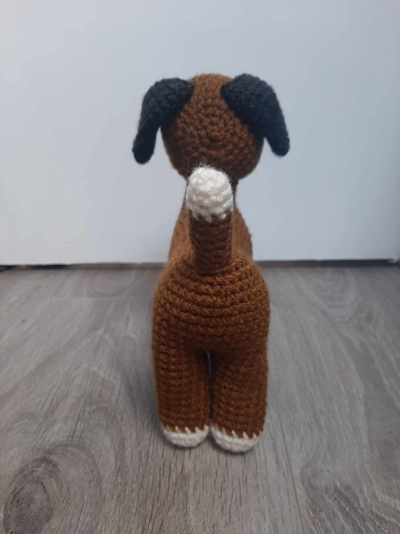 Op de afbeelding: Bruin gehaakt hondenspeelgoed met zwarte oren en witte voeten. De hond staat op een houten vloer.