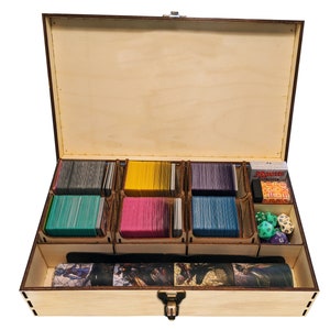 May include: A wooden card storage box with multiple compartments. The box contains stacks of colorful cards, dice, and a Magic: The Gathering card pack. The box is made of light-colored wood with a dark brown trim and a metal clasp.