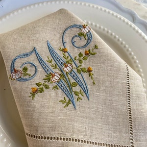 Peut inclure: Serviette en lin beige avec une broderie décorative de la lettre "W" en bleu clair, ornée de citrouilles et de feuillage d'automne. Posée sur une assiette blanche à bord perlé, idéale pour les tables d'automne.
