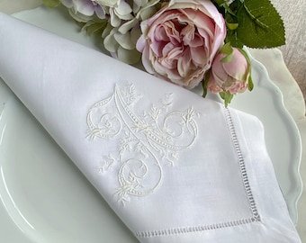 May include: White linen napkin with an embroidered letter "A" in an elegant floral design. The napkin is folded on a white plate, with pink and green flowers in the background. The napkin has a stitched border.