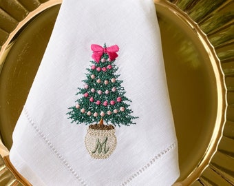 May include: White linen napkin with a stitched Christmas tree design. The tree is green with pink ornaments and a pink bow at the top. The tree sits in a pot with the letter "M". The napkin is folded on a gold plate.