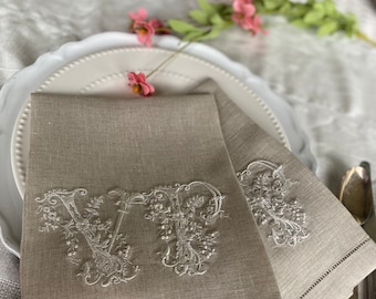 May include: Two beige linen napkins with embroidered initials, "VP", in a decorative floral script. The napkins are placed on a white plate, with pink flowers and silverware in the background. The napkins have a decorative stitched border.