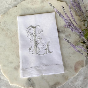 Può includere: Asciugamano in lino bianco con una lettera "I" ricamata in un design floreale decorativo. L'asciugamano è piegato e appoggiato su una superficie marmorea, con rametti di lavanda sullo sfondo. Il ricamo è di colore grigio chiaro.