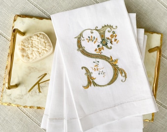 May include: A stack of white linen napkins embroidered with a gold and green floral "S" design. The napkins are on a marble tray with gold trim, next to a floral-shaped soap.